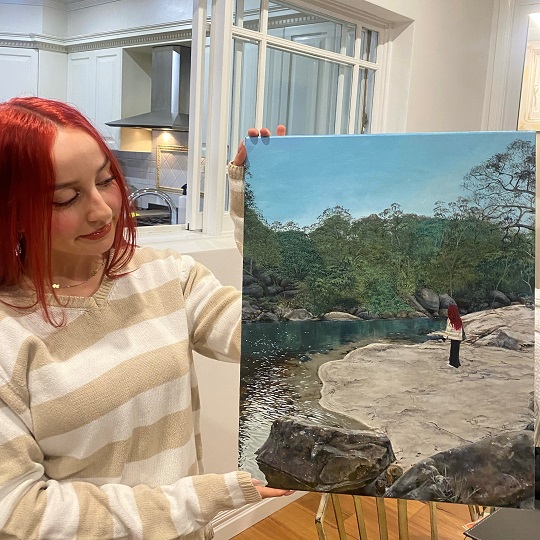 A photo of Mia holding up her entry artwork for the young creative awards, the painting of is a young woman standing on a rock formation that joins a stream with water, the background is green bush and blue sky, Mia wears a stripy white and coffee coloured top as does the woman in the painting.  Mia has long red hair and stands in a kitchen holding the artwork.